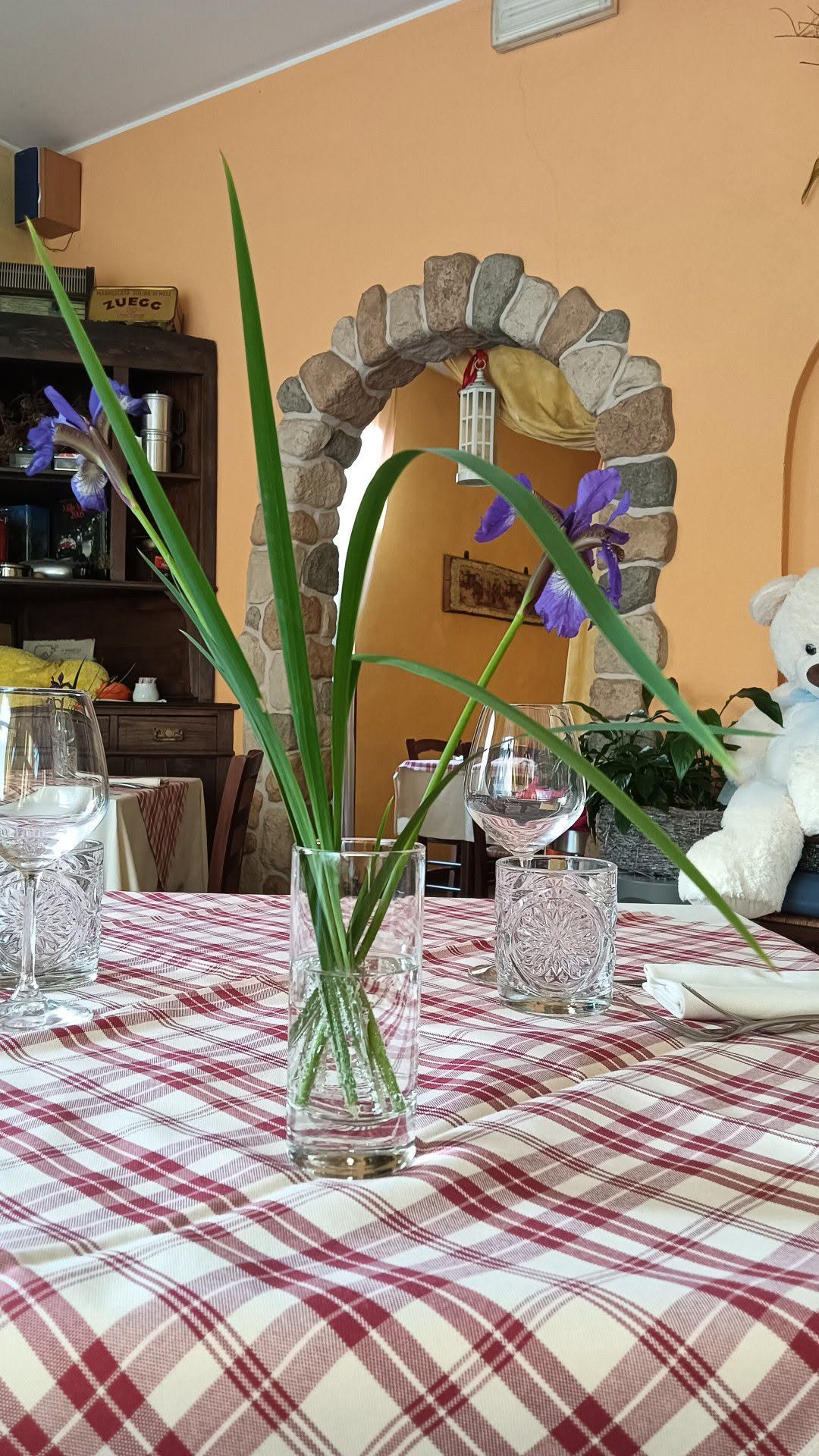 Tavolo con tovaglia a quadri bianchi e rossi, iris viola in un vaso di vetro e arco in pietra su parete arancione