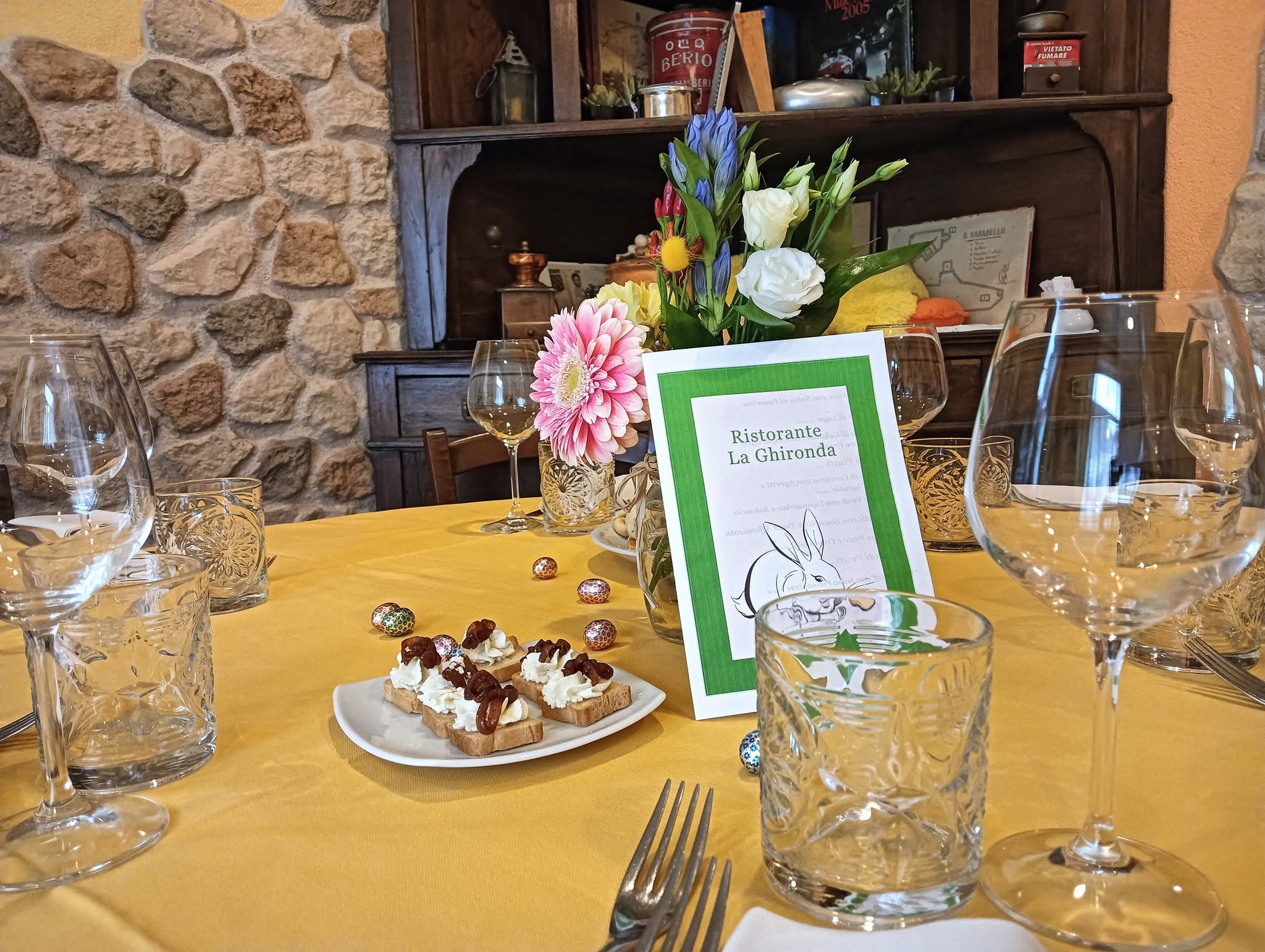Tavolo con tovaglia gialla, crostini con crema, ovetti di cioccolato, fiori e menù pasquale del Ristorante La Ghironda