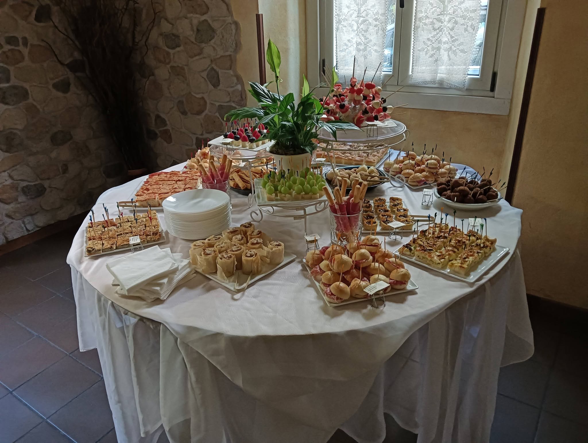 Buffet di stuzzichini su tavolo rotondo con tovaglia bianca, tramezzini, panini e spiedini di frutta, muro in pietra